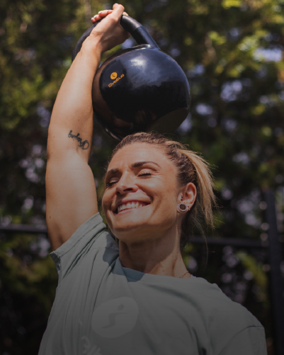 Une femme très souriante tenant un kettlebell olympique dans la main, bras tendu au dessus de la tête